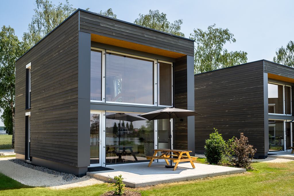 A modern chalet design features a large glass front showcasing the entrance and patio area. An outdoor table with benches is shaded by an umbrella, surrounded by green grass and landscaped shrubs, providing a spacious setting near the lake.