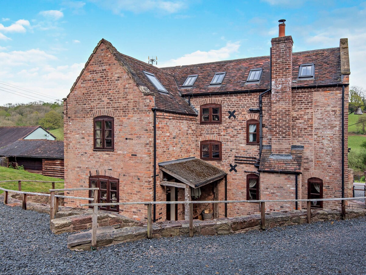 The exterior of the cottage is characterized by exposed brickwork and a traditional roof. Windows are present on multiple levels, allowing natural light into the property. A wooden porch provides an entryway, and gravel pathways lead to surrounding greenery.