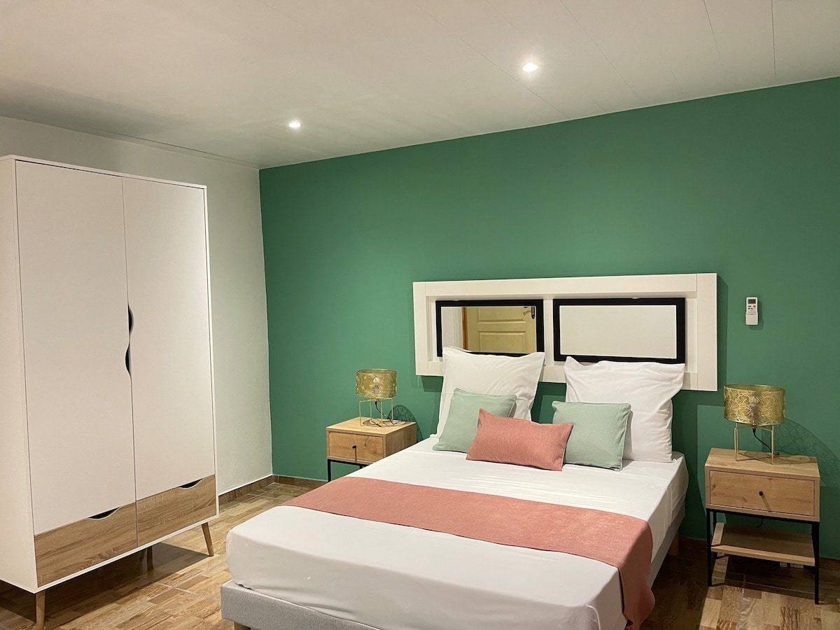 A spacious bedroom features a large bed dressed in soft linens, complemented by a decorative pink throw. Two bedside tables with lamps frame the bed, while an elegant wardrobe stands against the wall. A calming green accent wall adds a touch of tranquility.