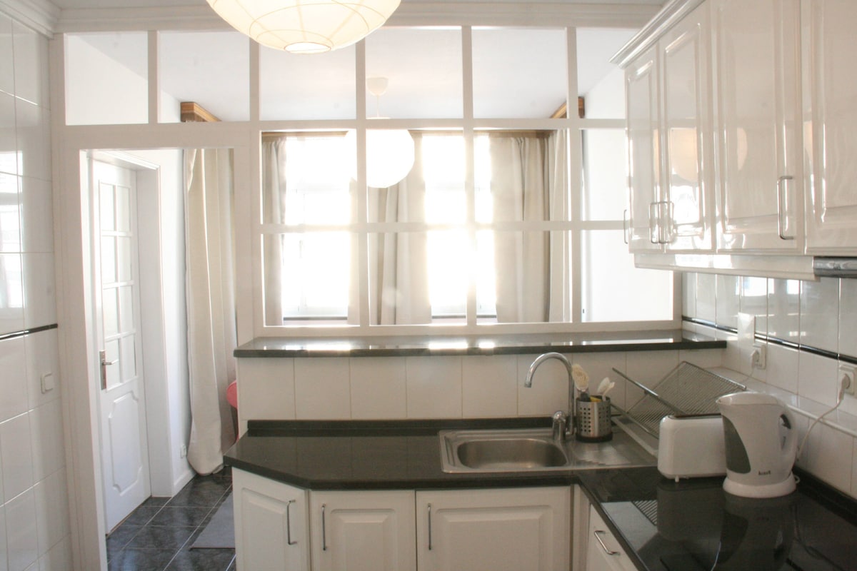 A bright kitchen area features white cabinetry and a sleek black countertop. A window allows natural light to fill the space, illuminating the room. The kitchen includes modern appliances, with a kettle and sink visible on the countertop.