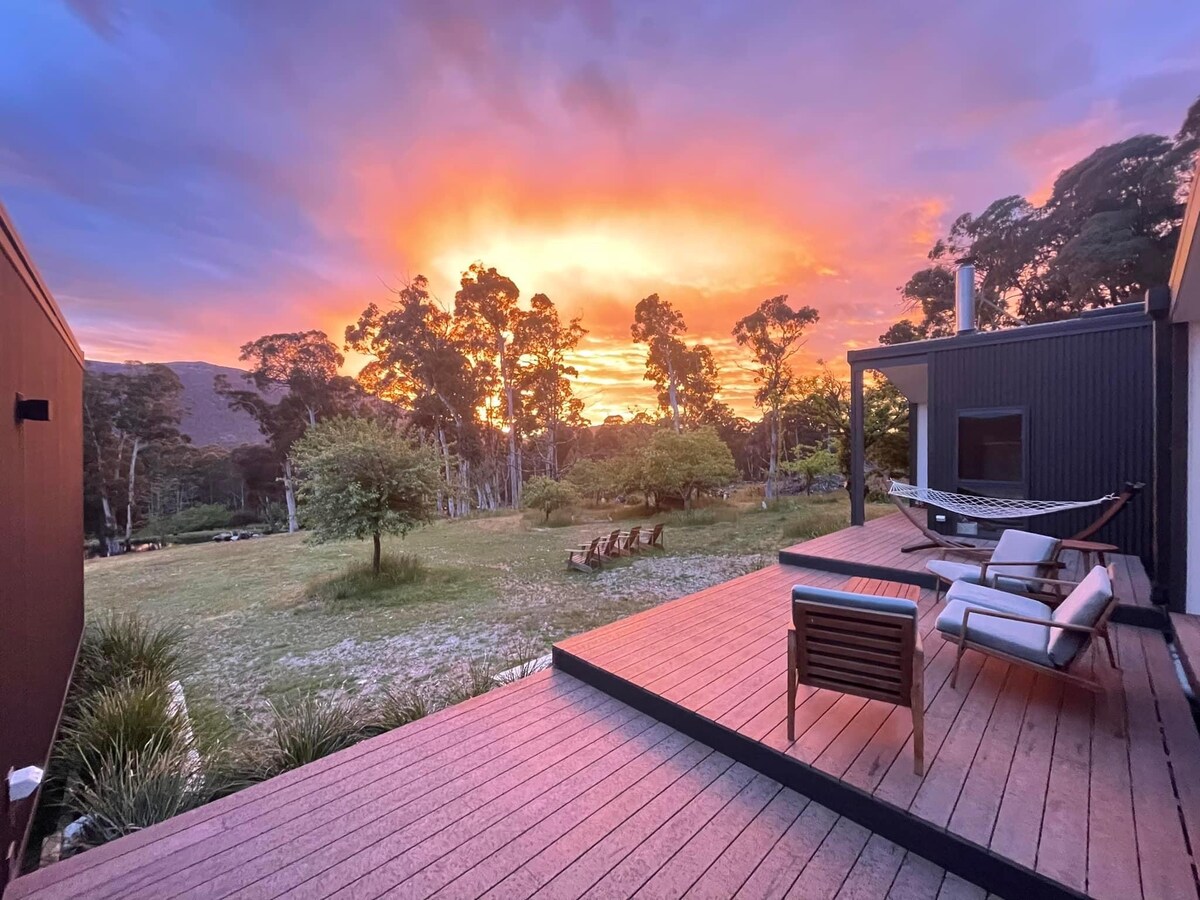 A serene outdoor deck features comfortable seating and a hammock, surrounded by lush greenery. A vibrant sunset casts warm hues across the sky, creating a tranquil backdrop for the landscape. The distant silhouettes of trees frame the scene, enhancing the natural beauty of the setting.