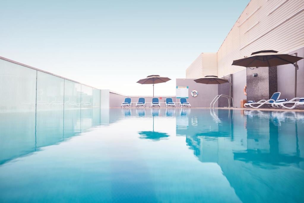 An outdoor swimming pool is pictured, featuring a clear blue surface that reflects the sky. Sun loungers with umbrellas are positioned around the pool, providing shaded areas for relaxation. The pool area is surrounded by a glass barrier, enhancing the serene atmosphere.