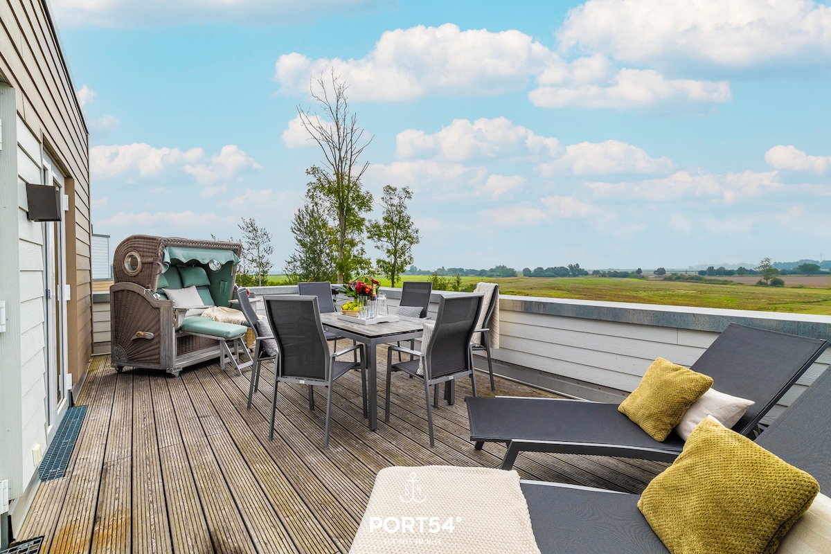 A spacious terrace features a dining table surrounded by comfortable chairs, along with lounge chairs positioned for relaxation. A cozy seating area includes a protective canopy. Lush green fields and a clear blue sky serve as a serene backdrop.