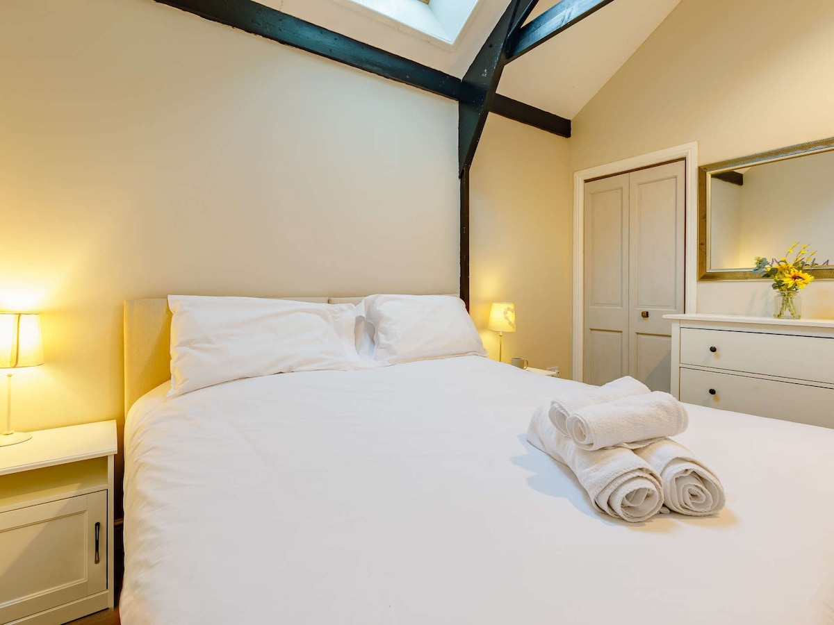 A bright bedroom features a spacious bed with crisp white linens and fluffy towels neatly arranged at the foot. Soft lamps provide gentle lighting, while a large skylight allows natural light to brighten the room. A dresser and mirror add functionality to the space.