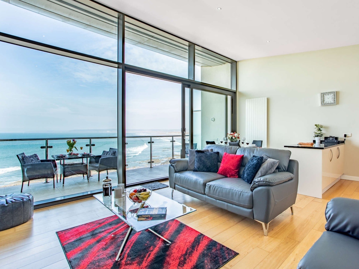 A spacious living area features a modern glass sliding door that leads to a private balcony with sea views. Light streams in, highlighting the contemporary furnishings, including a gray sofa and a coffee table with magazines. Vibrant red and black accents contrast against neutral tones.