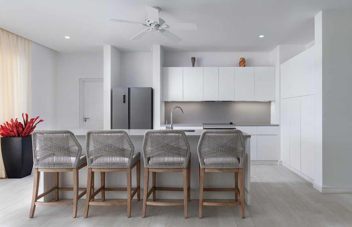 A modern kitchen features sleek white cabinetry and a large kitchen island with four woven bar stools. A stainless steel refrigerator and a well-appointed sink are present, along with minimalist décor. Natural light floods the space through sheer curtains, enhancing the open ambiance.