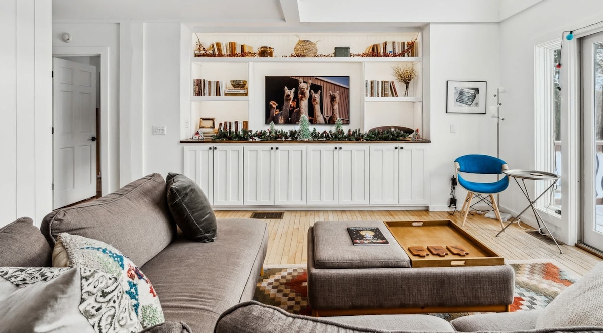 A cozy living area showcases plush, neutral-colored seating positioned around a central coffee table. A wall-mounted TV, framed by shelves filled with books and decor, stands as a focal point. Light streams in through large windows, enhancing the inviting ambiance.