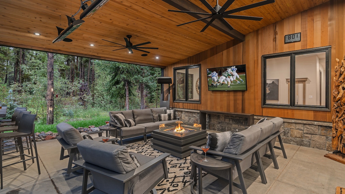 A covered outdoor seating area features a modern firepit surrounded by comfortable gray seating and two large ceiling fans. A television is mounted on the wall, and a bar with high stools is visible on one side, all set against a backdrop of lush greenery.