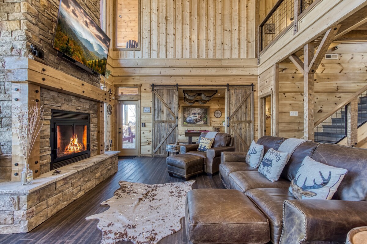 A spacious living area features a rustic stone fireplace, warmly lit by a fire. A large sectional sofa with accent pillows is arranged comfortably. Exposed wooden beams and walls create a cozy atmosphere, while a large television is mounted above the fireplace, enhancing the room's entertainment options.