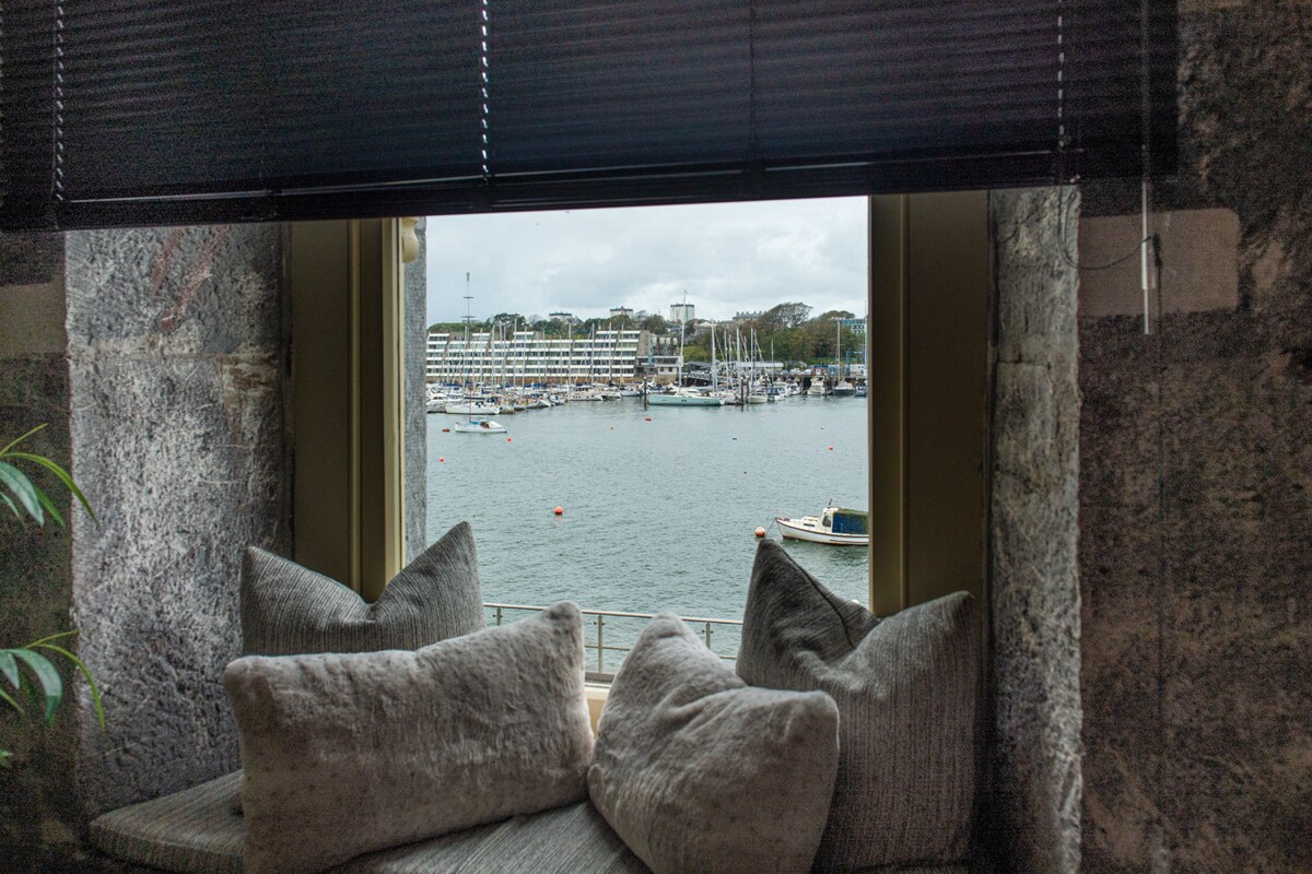 A comfortable window nook is adorned with soft cushions, offering a peaceful spot to relax. Outside, the view reveals boats gently bobbing on the water, with a marina and trees visible in the background under a cloudy sky.