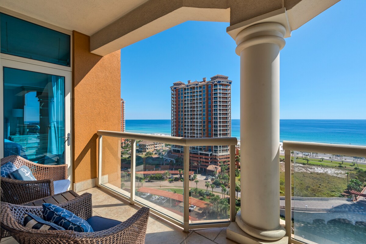 A spacious balcony features comfortable wicker chairs, providing a relaxing spot to enjoy expansive views of the Gulf of Mexico and nearby beach. Nearby towers reflect a warm sunlit ambiance, while the clear blue sky enhances the coastal atmosphere.
