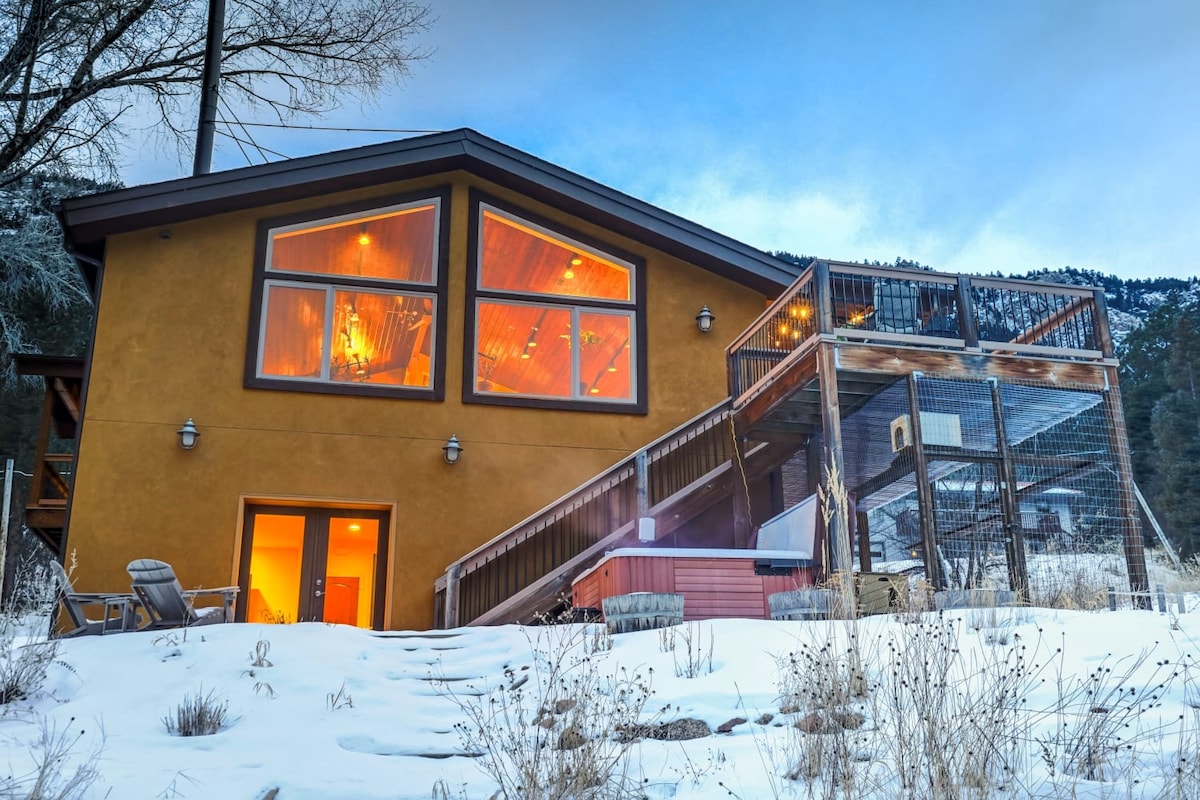 The exterior of a mountain retreat showcases a warm-toned building with large windows reflecting soft light. Steps lead up to a deck featuring a hot tub, surrounded by snow and gentle landscaping. The backdrop consists of trees and mountains, creating a serene environment.