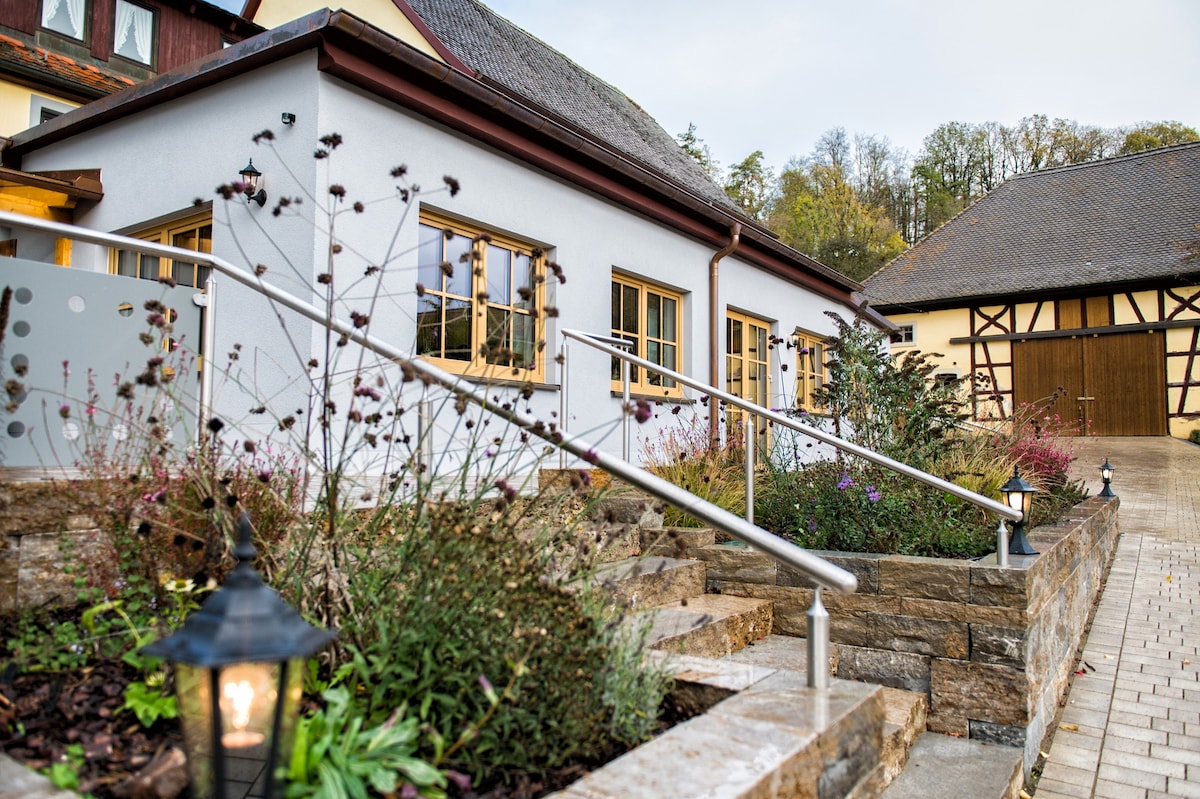 The exterior of the building features a welcoming approach with stone steps leading to the entrance. Several wooden-framed windows are visible, flanked by a well-maintained garden with various plants and flowers. Decorative lanterns line the path, providing additional ambiance.