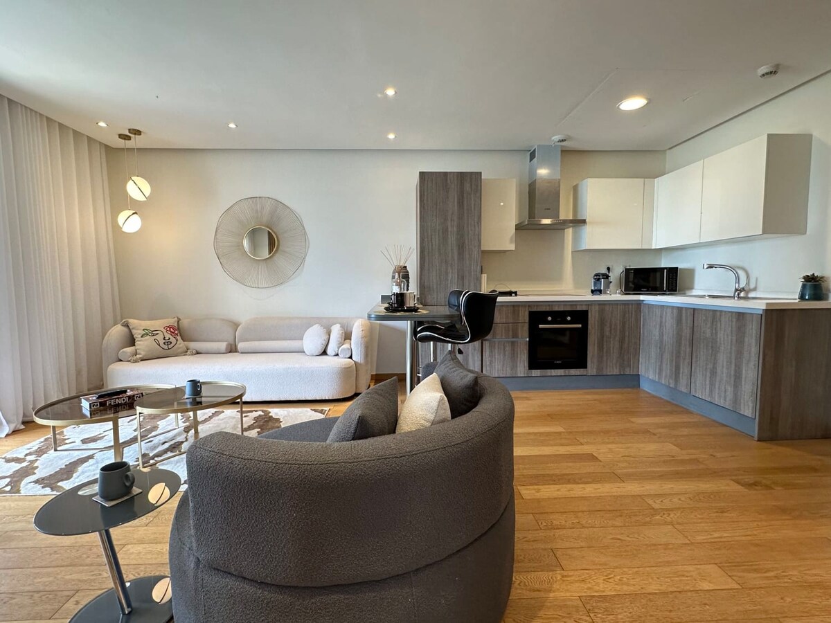 A contemporary living area features a light-colored sofa accompanied by circular cushions. A round mirror hangs above a minimalist coffee table. The gray armchair offers a comfortable seating option. Modern kitchen appliances are visible in the background, complemented by wooden cabinetry.