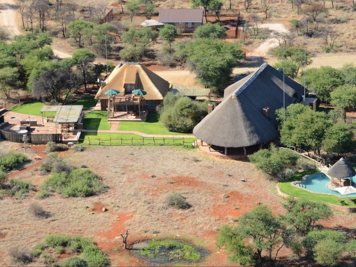 An aerial view captures various outdoor structures, including thatched-roof buildings surrounded by greenery. A pool glimmers nearby, complemented by landscaped lawns. Paths and natural terrain form the backdrop, enhancing the sense of space and serenity.