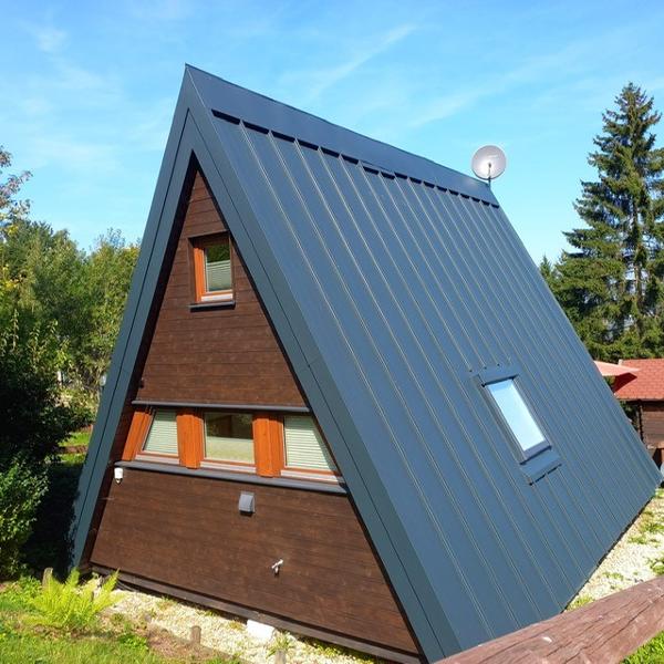 The chalet features an A-frame design with a dark wooden exterior and a steel grey roof. Two rectangular windows are framed by orange shutters, and a satellite dish is mounted on the roof. Lush greenery surrounds the structure, enhancing the natural setting.