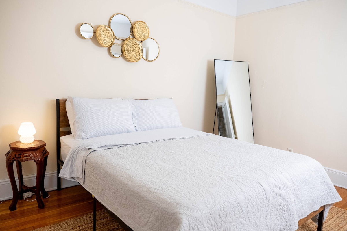 A serene bedroom features a spacious bed covered in a light quilt. A round mirror collection adorns the wall above the bed, while a full-length mirror stands in the corner. Soft lighting is provided by a small lamp on a decorative table.