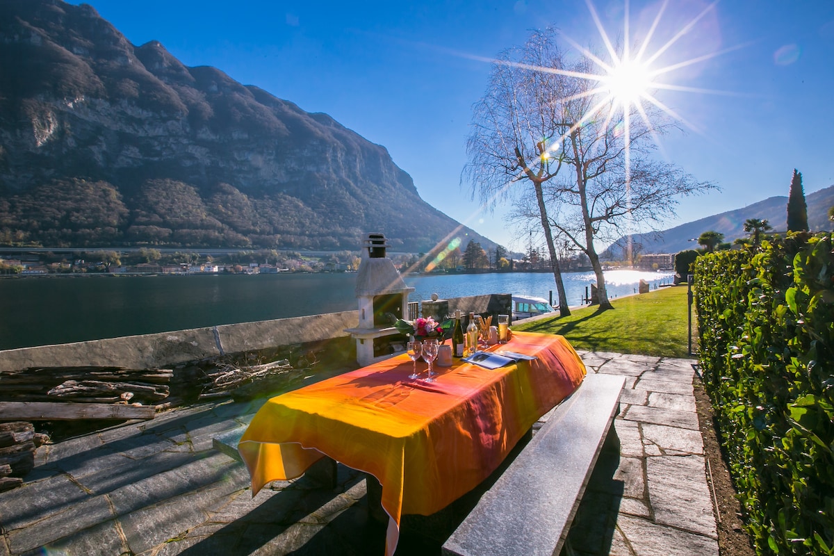 An outdoor dining area is set with a vibrant tablecloth, featuring a table surrounded by chairs. The scenic backdrop showcases the lake and mountains under a clear sky, with sunlight reflecting off the water. A barbecue facility is positioned nearby.