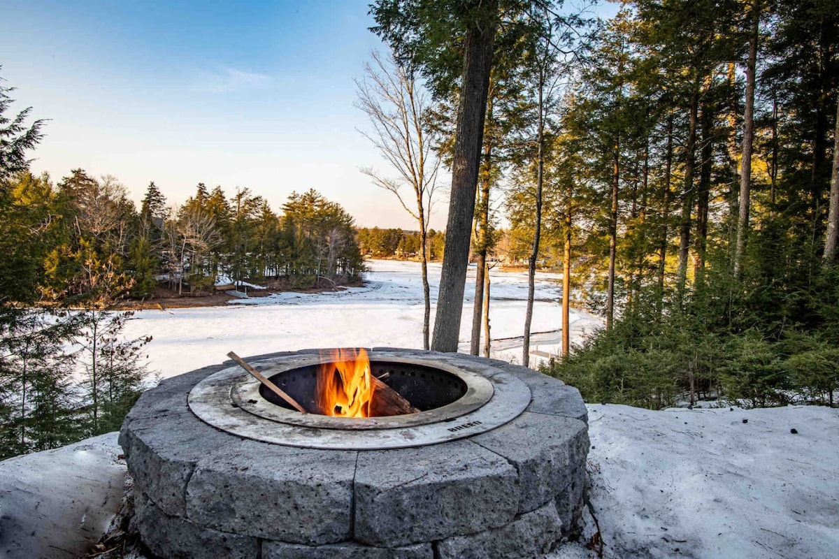 A circular stone fire pit is surrounded by snow and trees, offering a warm gathering spot. A small flame flickers within the pit, creating a cozy atmosphere against the backdrop of a serene, icy landscape visible in the distance.