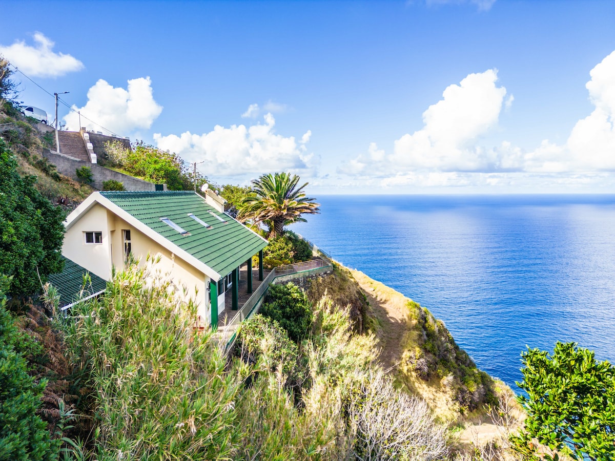The villa is positioned on a cliffside, surrounded by lush vegetation and offering panoramic views of the Atlantic Ocean. The bright exterior features a green roof and large windows, allowing natural light to fill the living spaces. A clear blue sky with scattered clouds enhances the serene setting.