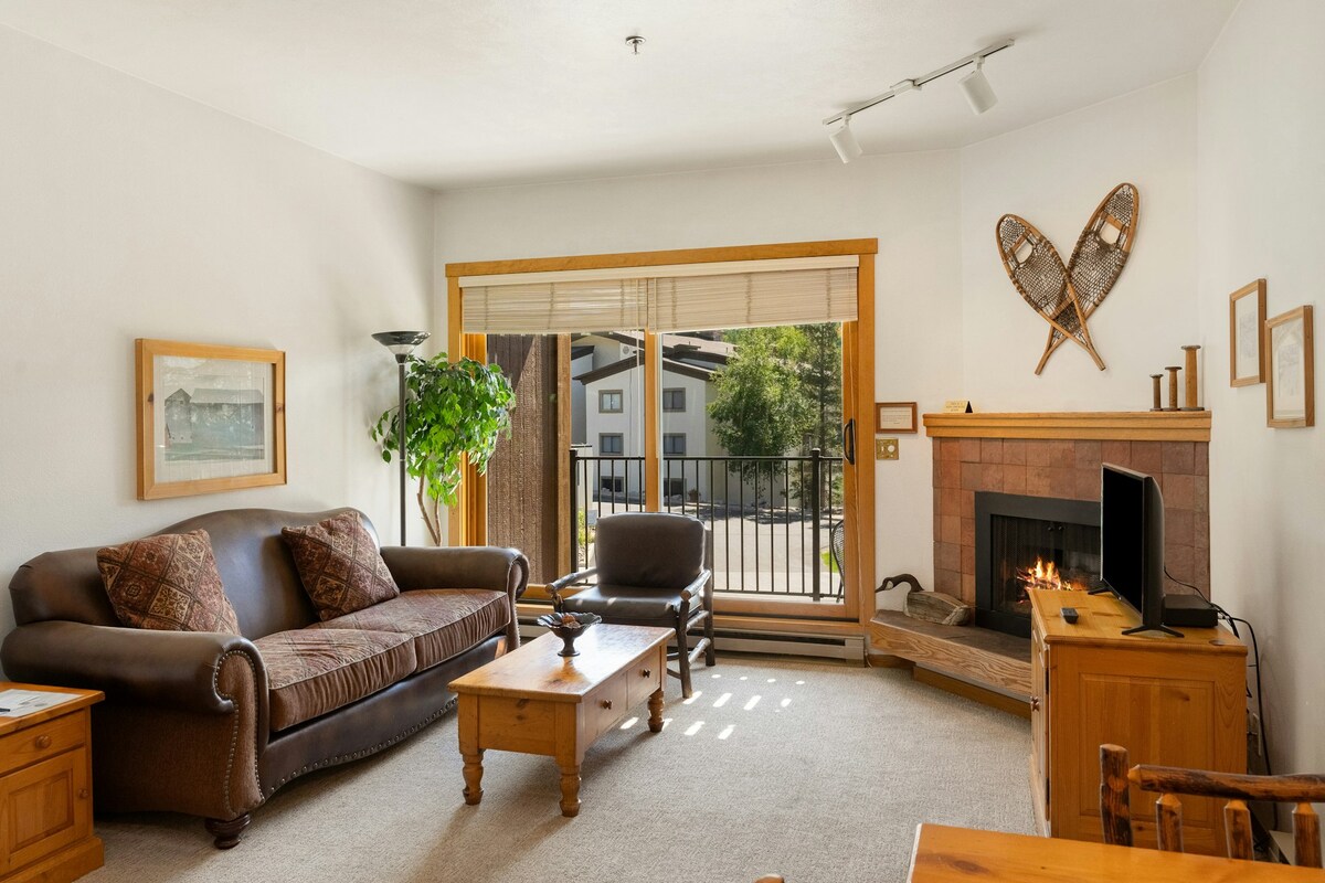 A cozy living area is presented with a comfortable brown sofa and armchair, facing a gas fireplace set within a stone hearth. Natural light fills the space through sliding glass doors, which open to a small balcony. Wall art and a decorative plant add a hint of warmth.
