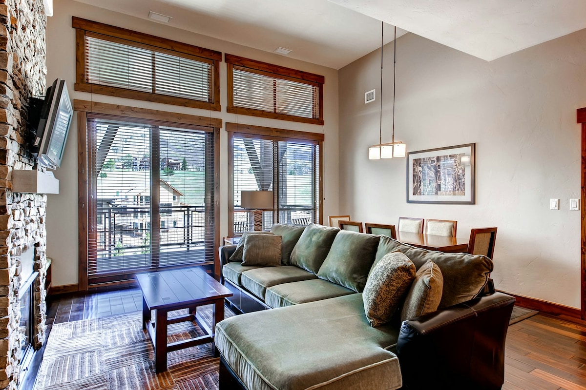 A spacious living area is highlighted by large windows, allowing natural light to fill the room. A comfortable sectional sofa faces a flat-screen television mounted above a stone fireplace. A dining table for six is positioned nearby, with warm wooden accents throughout the space.