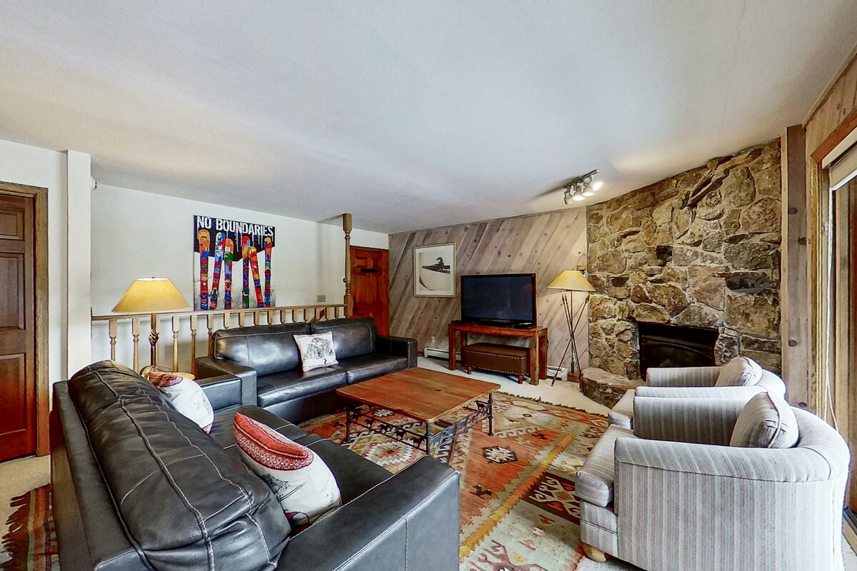 A spacious living area features dark leather sofas and two upholstered chairs arranged around a wooden coffee table. A stone fireplace with a rock facade is positioned in the corner, with a TV mounted nearby. Natural light filters in through sliding glass doors.