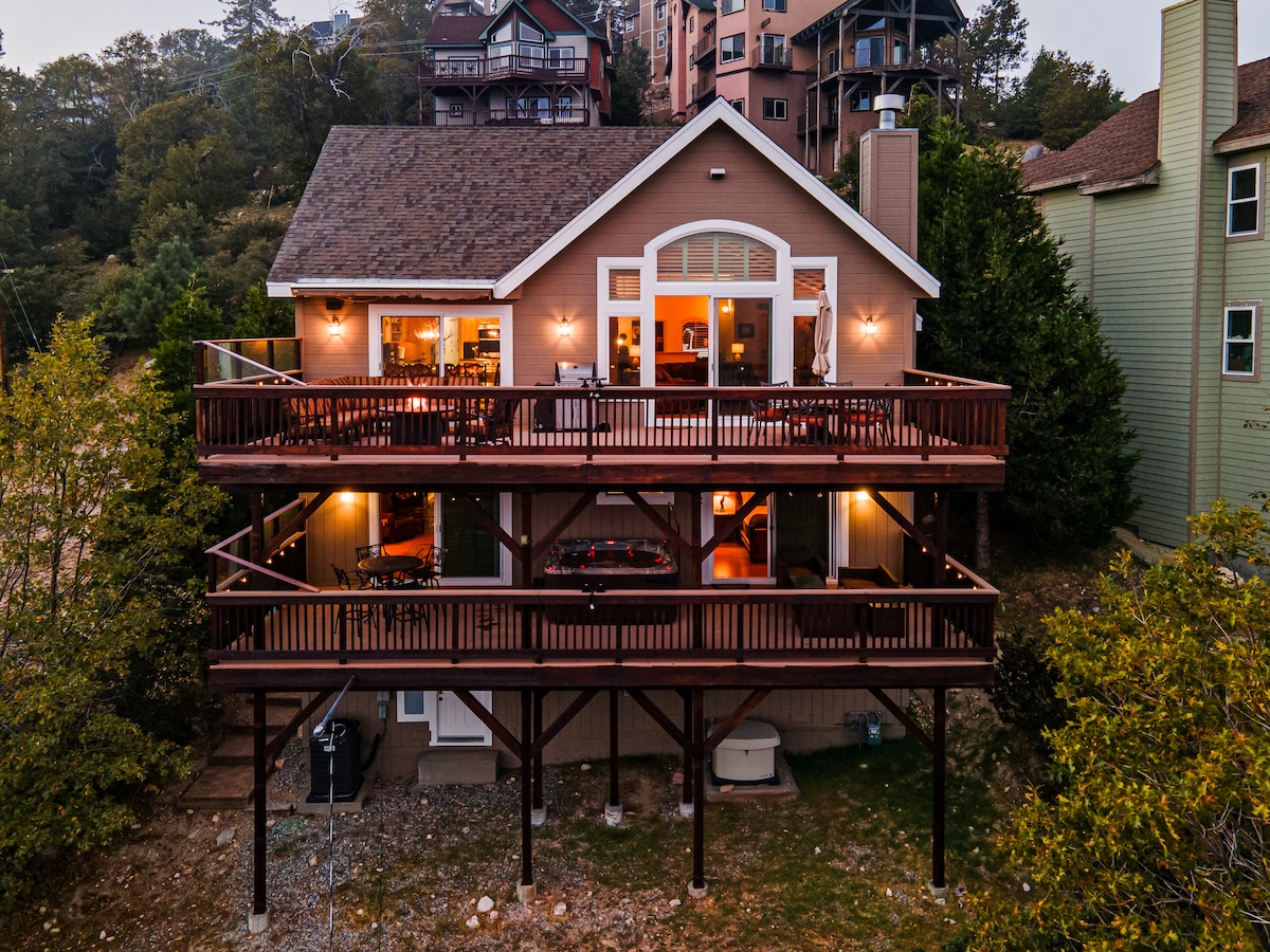 The exterior of the lodge showcases an inviting facade with warm lighting illuminating multiple dining and lounging areas on expansive wooden balconies. The structure is elevated slightly above ground, surrounded by greenery and neighboring homes, creating a peaceful retreat.