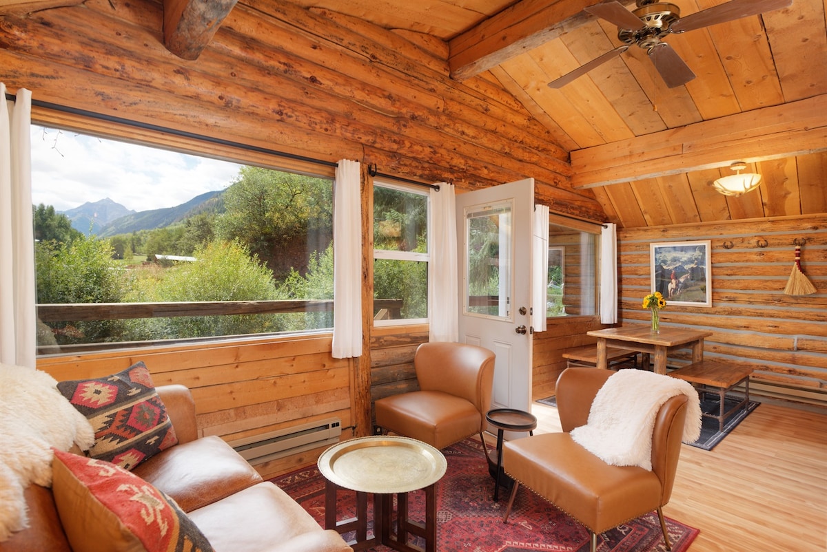 The interior of the cabin features rustic wooden walls and ceiling, complemented by large windows offering views of the surrounding landscape. Comfortable seating arrangements include two armchairs, a small coffee table, and a dining table in the background, creating a cozy gathering space.