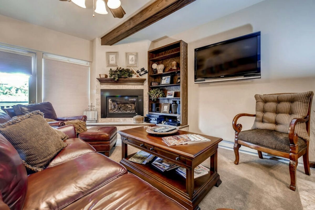 A comfortable living space features a rich leather sofa with accent cushions and a wooden coffee table displaying magazines. A gas fireplace is nestled between built-in shelves filled with decorative items. A flatscreen TV is mounted above the fireplace, and natural light enters through nearby windows.