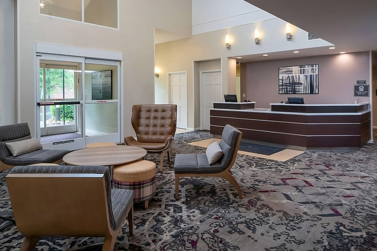 The lobby area features a modern reception desk and an inviting seating arrangement. Comfortable armchairs and a low round table are surrounded by decorative carpet. Natural light filters through the glass entrance, highlighting the contemporary decor and warm color palette.