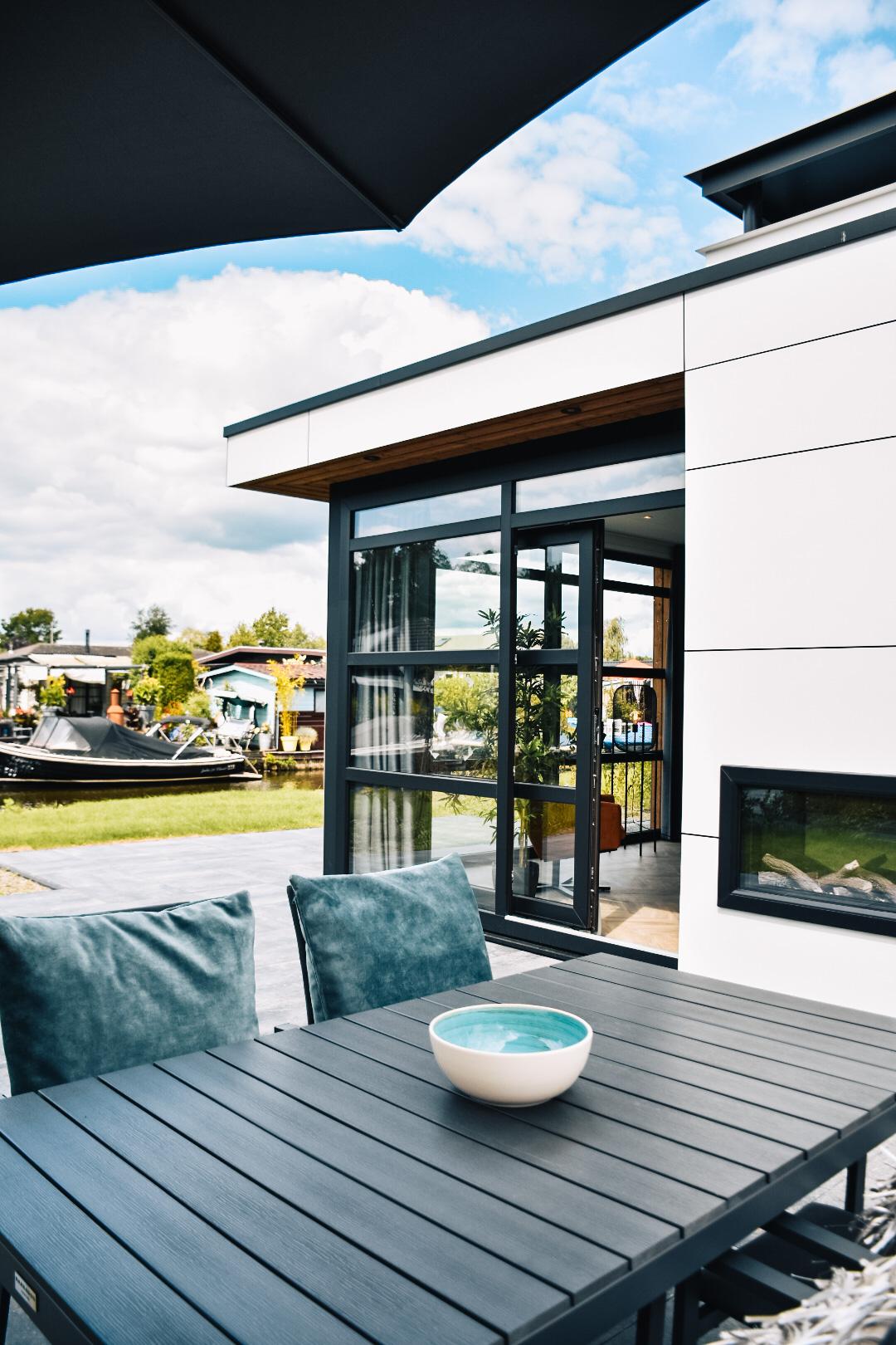 A modern outdoor seating area features a sleek black table accompanied by two cushioned chairs. A bowl rests on the table, contrasting with the surrounding green landscape and serene water views. Large glass doors provide access to the interior space while enhancing natural light.