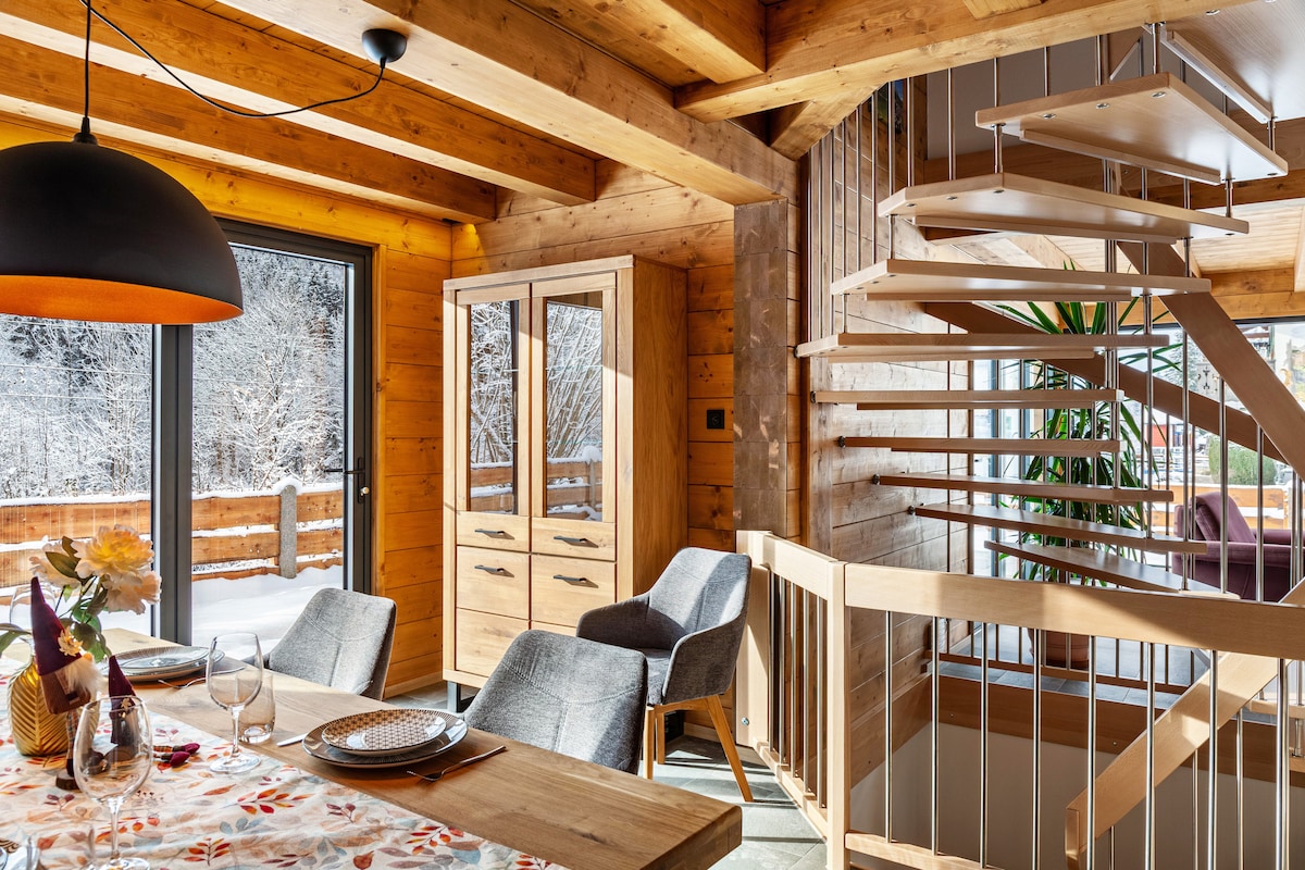A bright dining area is featured with a wooden table set for a meal. Soft grey chairs surround the table, while a stylish cabinet provides storage. A spiral staircase leads to the upper level, complemented by large windows that introduce natural light and views of the snowy outdoors.