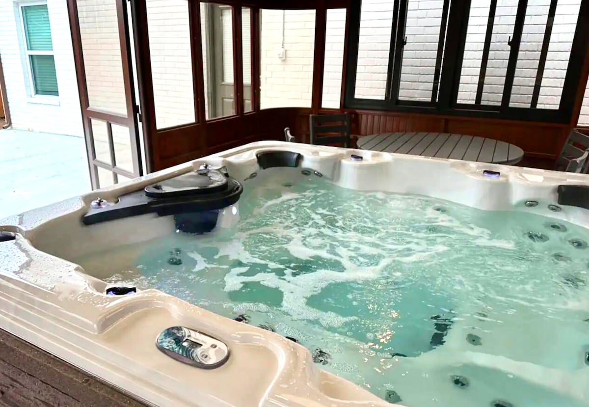 A private enclosed area features a spacious hot tub with multiple jets and clear, bubbling water. The surrounding walls are primarily glass, allowing natural light to illuminate the space. Outdoor seating and a table are available for relaxation and socializing.