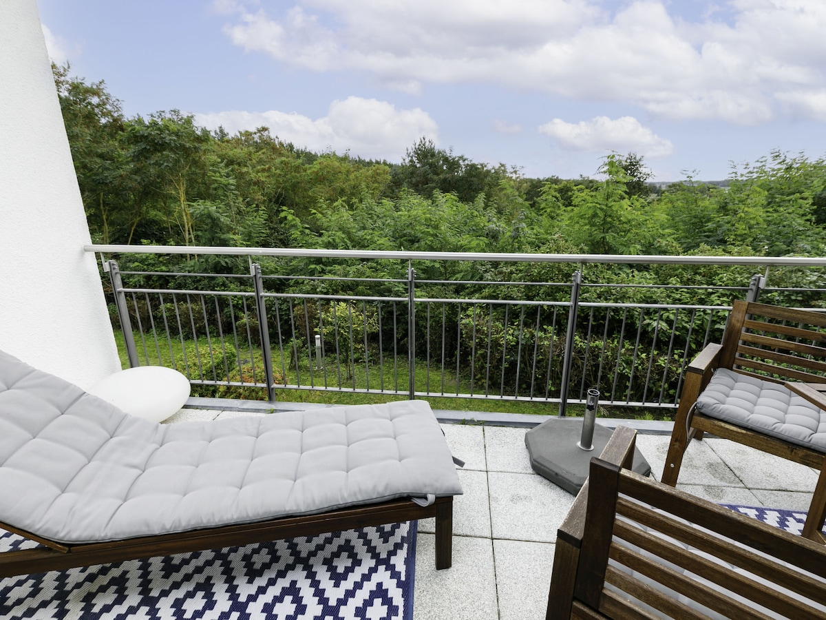 A balcony area features a lounge chair and wooden seating set, positioned on a patterned rug. Lush greenery is visible in the background, providing a natural setting. The balustrade offers unobstructed views of the surrounding landscape.