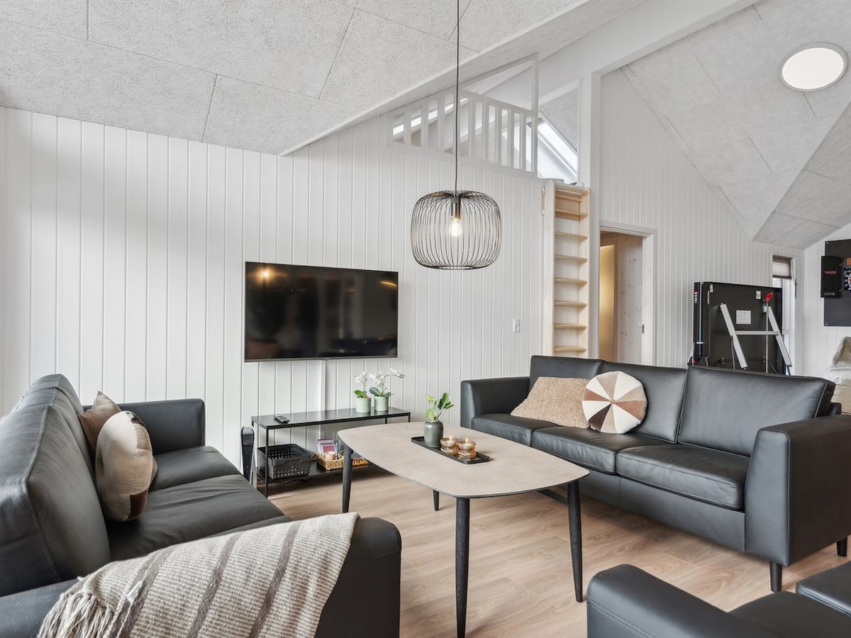 The living room features dark leather sofas arranged around a light wooden coffee table. A television is mounted on the wall across from the seating area, and a decorative pendant light hangs above. Light-colored walls are complemented by natural sunlight entering from a nearby window.