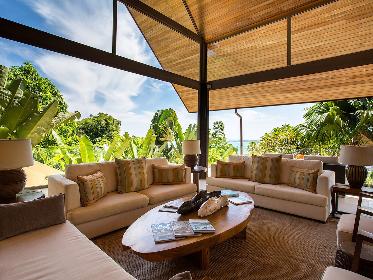 The open-plan living area features large glass doors that invite natural light and views of the surrounding greenery. Soft sofas are arranged around a wooden coffee table, complemented by warm lighting from side lamps. The high ceiling adds to the spacious feel of the room.