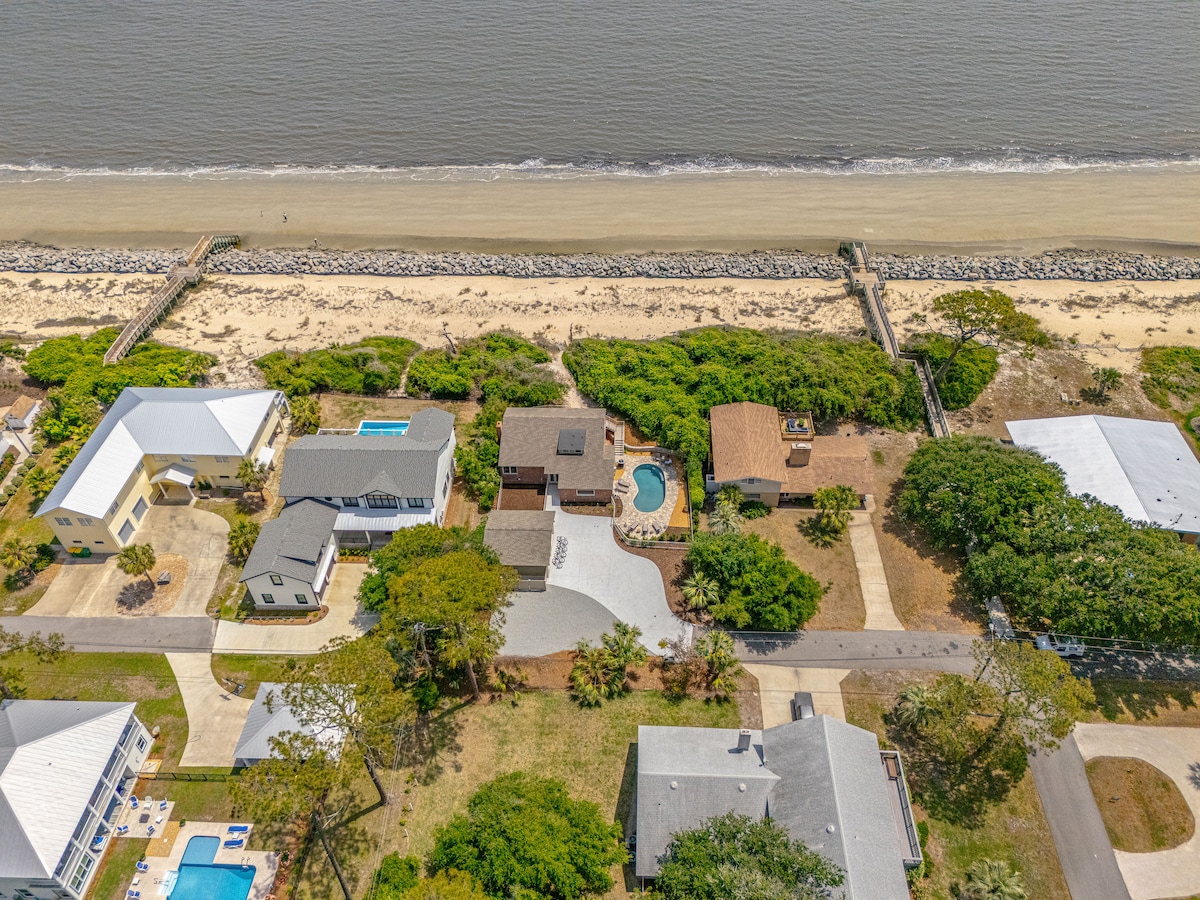 An aerial view captures the coastal setting, highlighting the home's proximity to the shoreline. The surrounding landscape features a mix of greenery and nearby residences, with beach access visible along the sandy shore. A pool is also noted on the property.