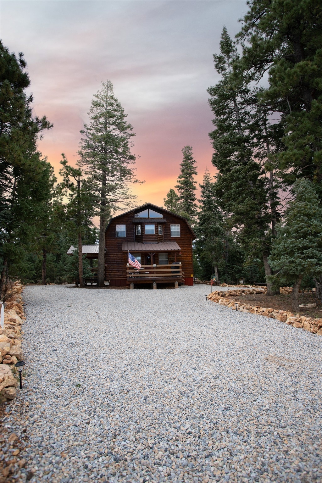 A rustic cabin stands surrounded by tall pine trees, featuring a welcoming porch adorned with an American flag. The gravel driveway leads up to the structure, framed by natural stone edging. A colorful sunset sky casts a warm glow over the scene.