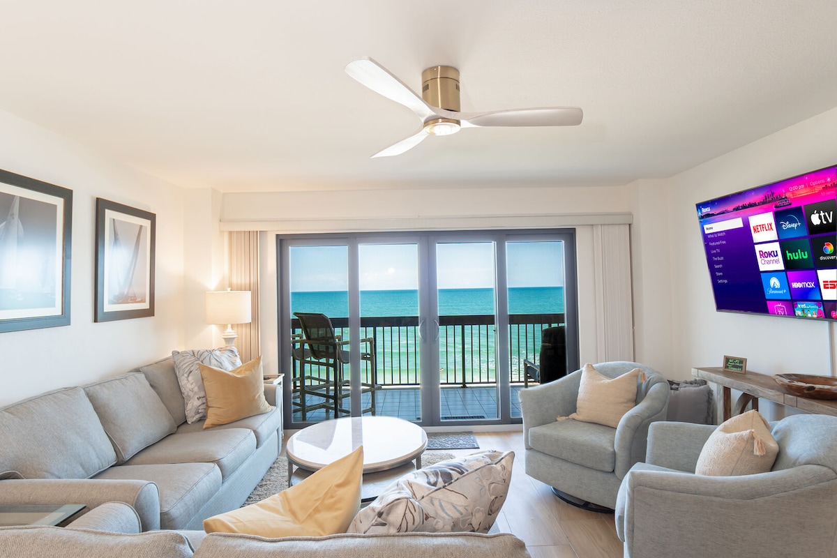 A welcoming living area features light grey sofas and a round coffee table. A ceiling fan is present, and a large television displays streaming options. Floor-to-ceiling glass doors provide an unobstructed ocean view, enhancing the bright and airy atmosphere of the space.