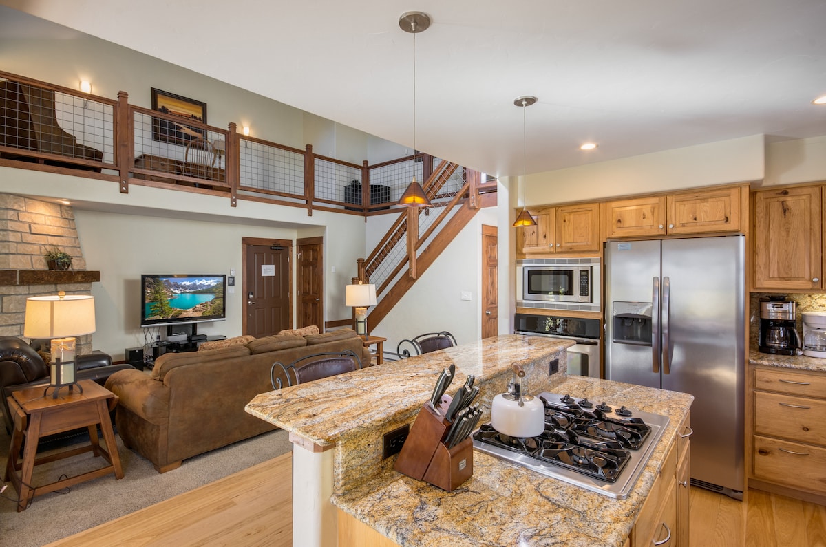 A well-appointed kitchen is visible with a spacious island featuring a gas cooktop and contemporary appliances. A comfortable living area is seen in the background, including a sofa and wall-mounted television. Natural light is provided by multiple windows, enhancing the inviting space.