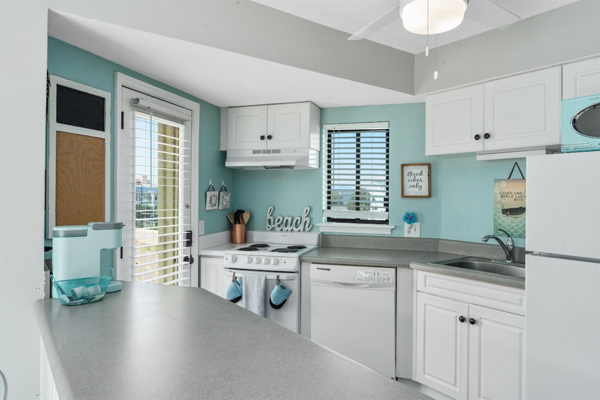 The kitchen is designed with a light, coastal color scheme featuring aqua walls and white cabinetry. Countertops provide ample workspace, complemented by modern appliances, including a refrigerator and dishwasher. Natural light enters through a window, while decorative elements emphasize the beach theme.