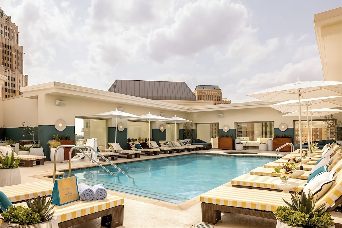 The outdoor swimming pool area is surrounded by lounge chairs and umbrellas, offering a relaxing space for guests. Potted plants add greenery, while the building's architecture lends a modern touch. Large windows can be seen in the background, creating a light-filled environment.
