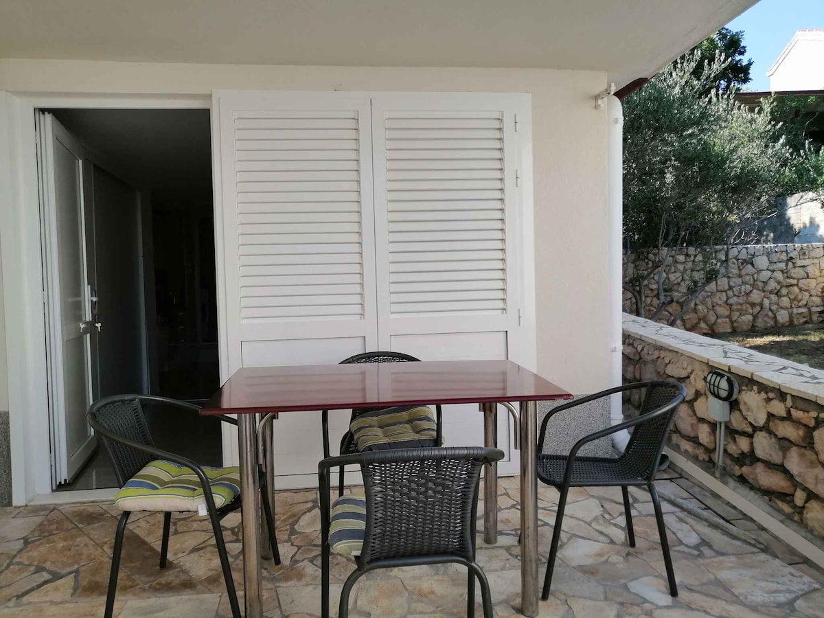 A terrace area is depicted, featuring a small table and four chairs set on a stone floor. The area is enclosed by a low stone wall and surrounded by greenery, with the entry to the studio visible in the background.