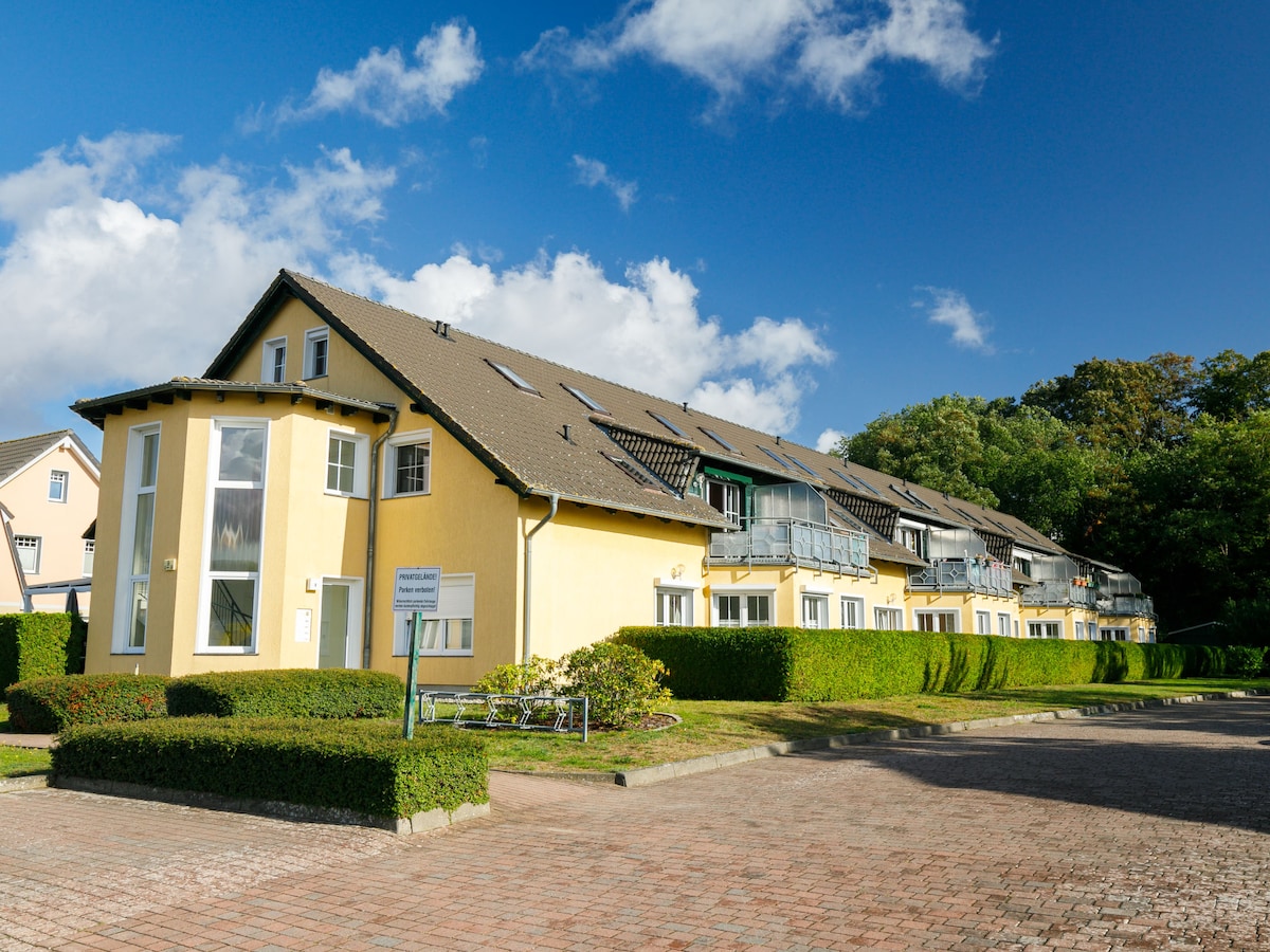 The exterior of a two-story apartment block is visible, featuring a charming yellow façade and a landscaped area with neatly trimmed hedges. Several balconies can be seen on the upper level, under a clear blue sky with scattered clouds.
