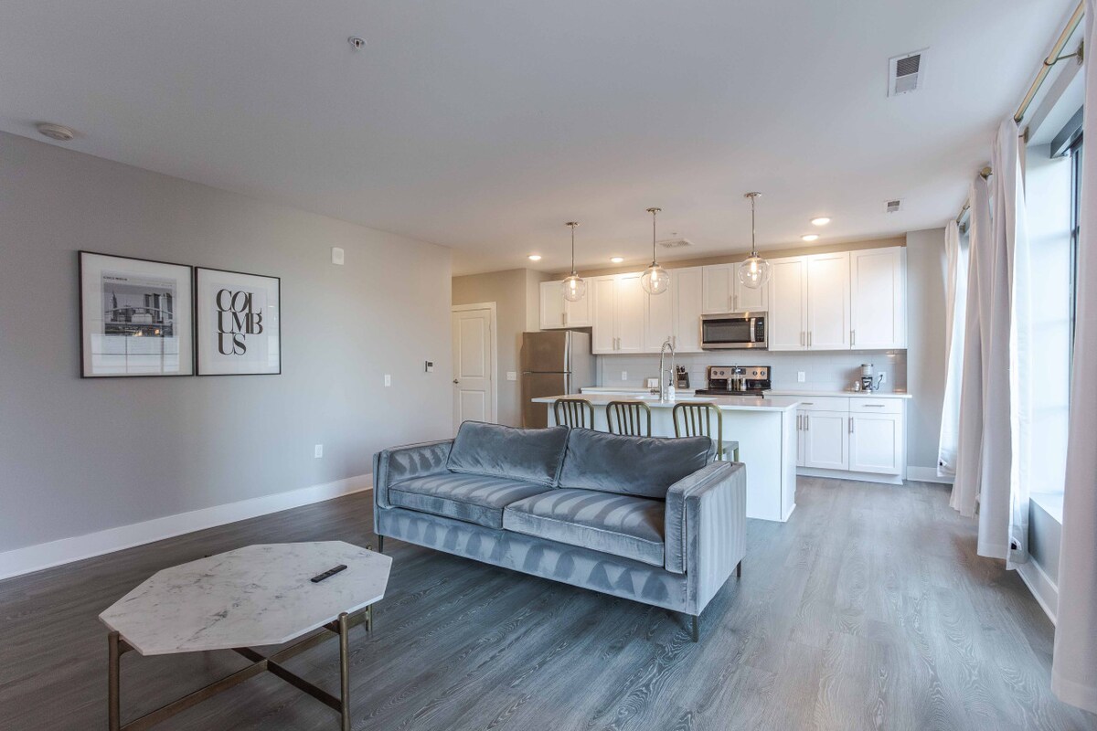 An open living space is presented, featuring a cozy sofa facing a modern kitchen with stainless steel appliances. A stylish coffee table sits in front of the sofa, while large windows allow ample natural light to illuminate the neutral color palette.