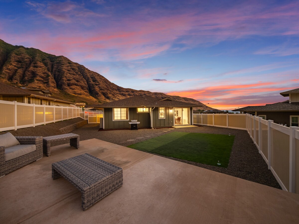The outdoor patio features a seating area with modern wicker furniture, arranged on a concrete surface, overlooking a grassy lawn. The backdrop displays dramatic mountains beneath a vibrant sky at dusk, enhancing the serene atmosphere of the space.