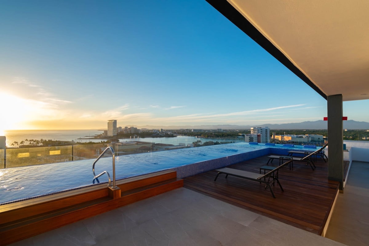 An expansive rooftop terrace features an infinity pool overlooking a scenic view of the coastline at sunset. Relaxing lounge chairs are positioned nearby, creating an inviting space for enjoyment while taking in the surrounding landscape.