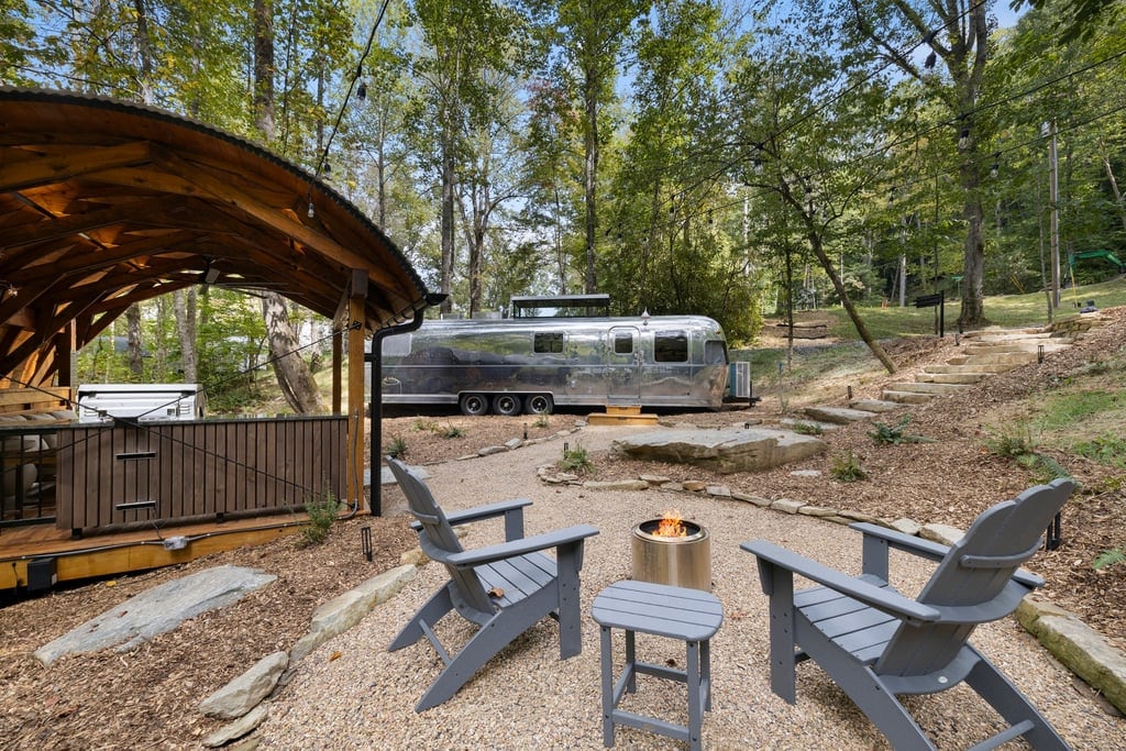 An inviting outdoor space features two adirondack chairs placed around a firepit, surrounded by a gravel pathway. A covered deck area and the sleek exterior of the Airstream are visible in the background, framed by lush trees and natural landscaping.
