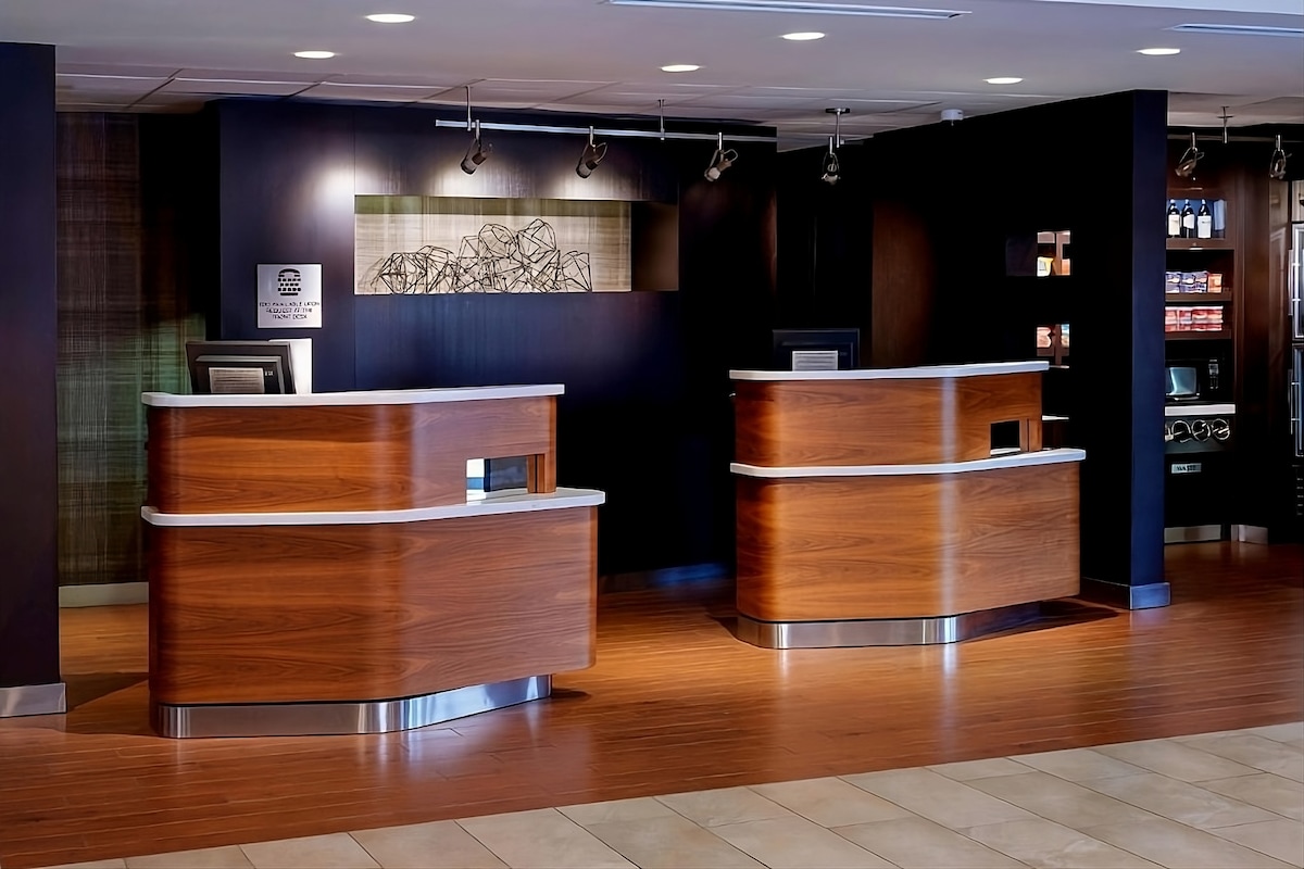 The reception area is characterized by two wooden service desks with smooth finishes. A minimalist artwork is displayed on the wall behind, contributing to a modern aesthetic. The flooring is light-colored, complementing the warm tones of the wood.